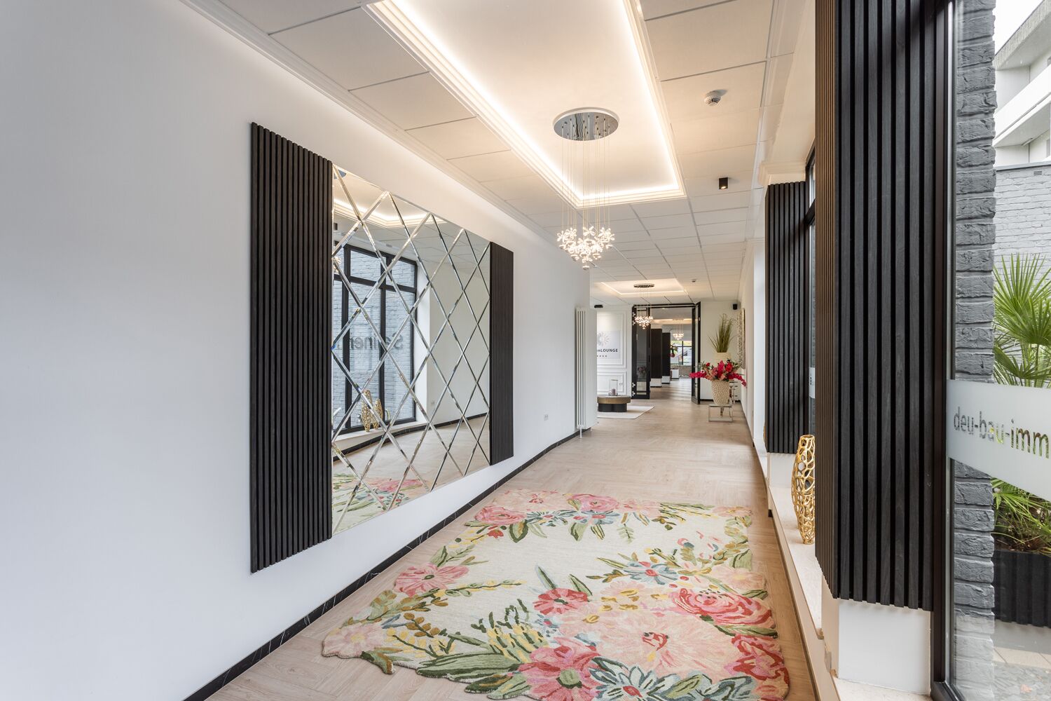 Un pasillo moderno con alfombra floral, una araña y vista a un área de descanso.