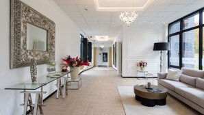 A spacious living room with a glass coffee table, a sofa, and a chandelier. There is a dining area with a glass table and chairs, and a large mirror on the wall.