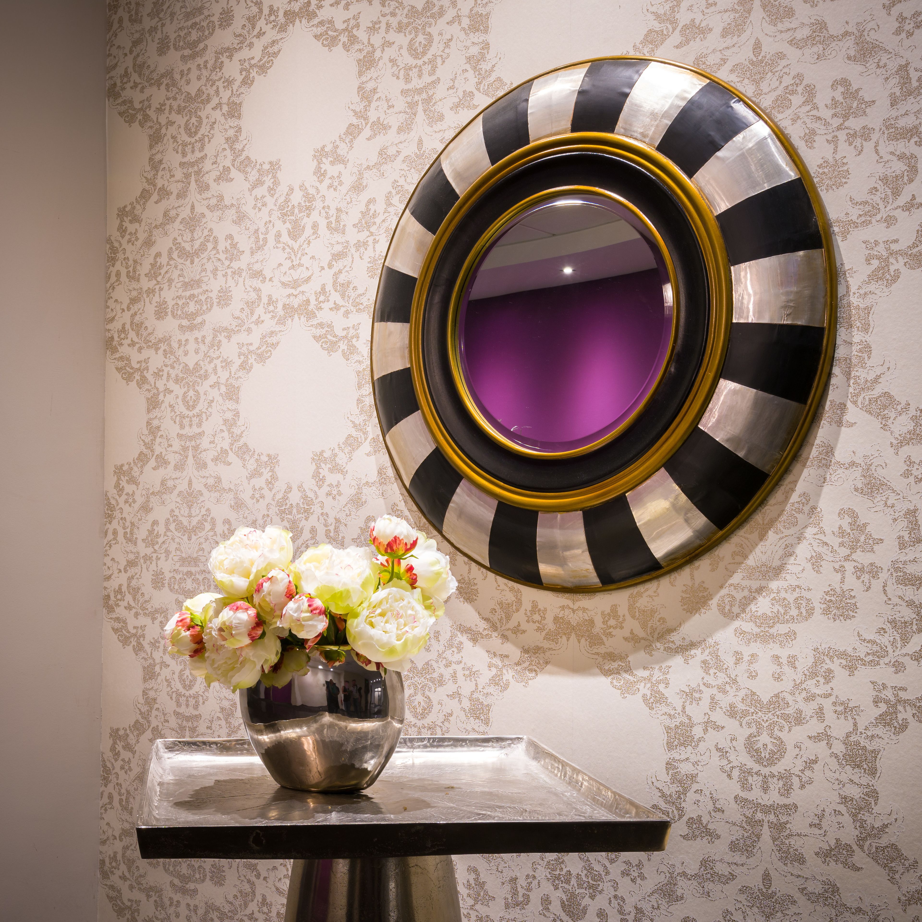 A mirror with a black and white striped frame, a vase with flowers, and a patterned wallpaper.