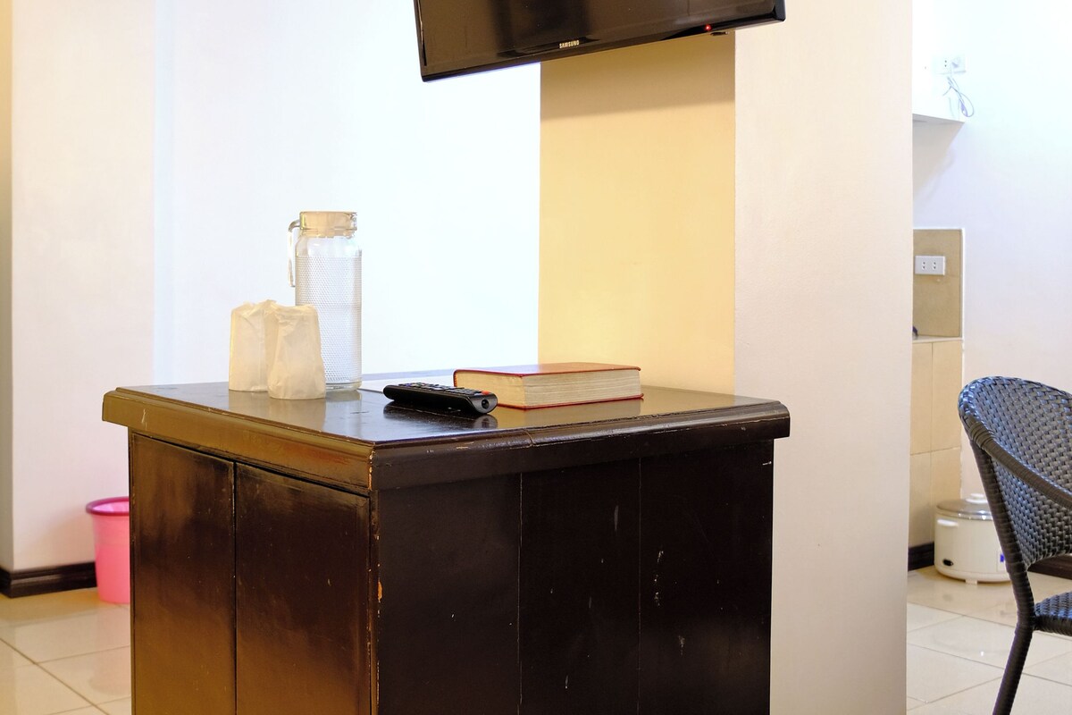 A room with a flat-screen TV mounted on the wall, a small wooden desk with a book and remote control, a trash bin, and a chair.