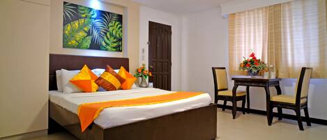 A hotel room with a bed, a small table with chairs, a door, and a painting on the wall.