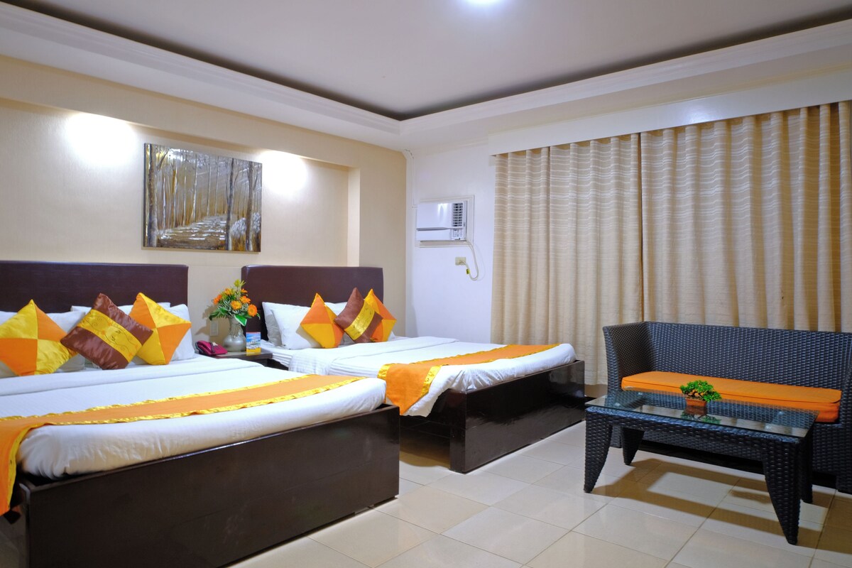 A hotel room with two beds, a sofa, a small table, a painting on the wall, and an air conditioning unit.