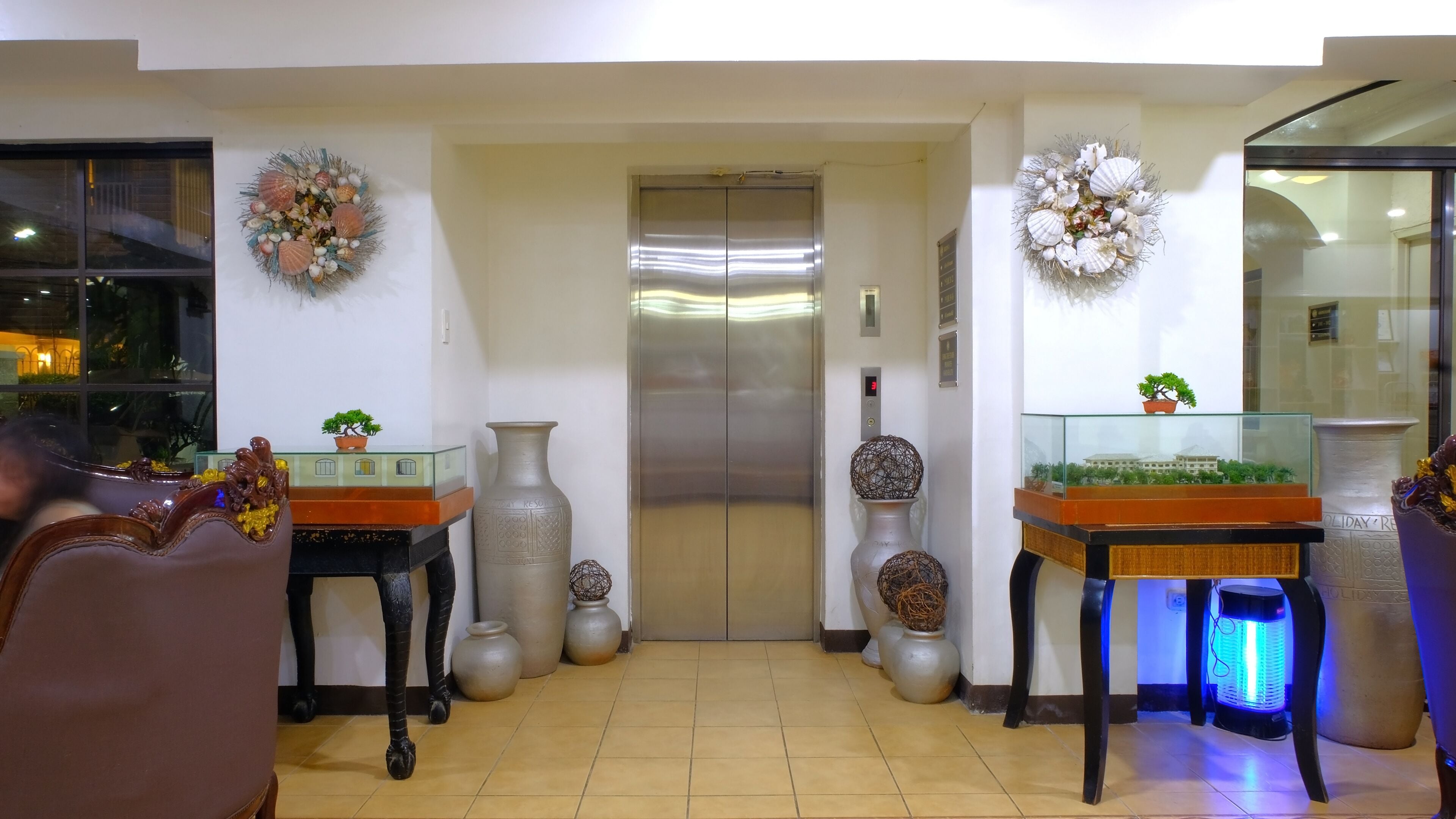 A lobby with an elevator, two tables with decorative items, and a glass display case with a plant.