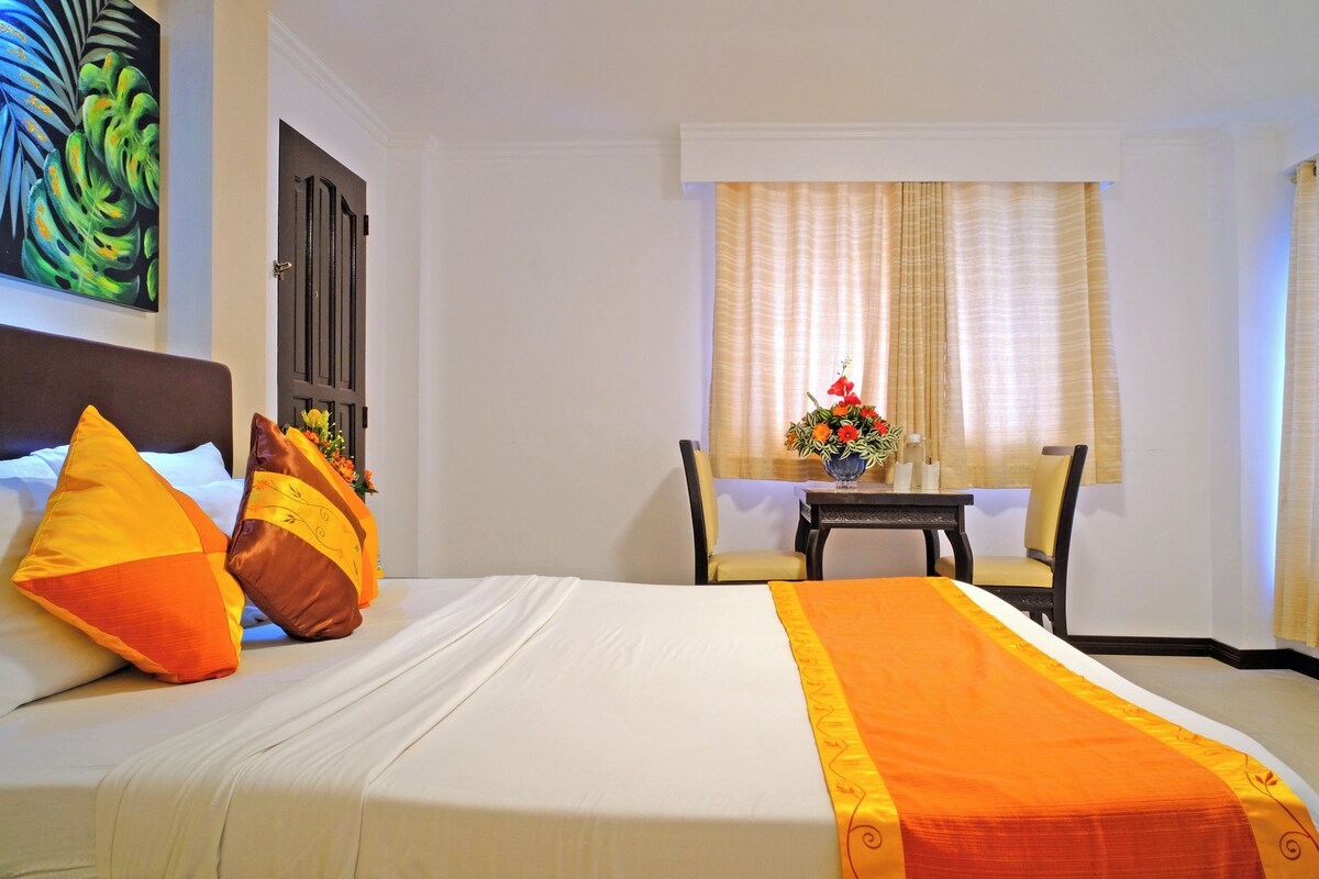 A hotel room with a bed, pillows, a table with a vase of flowers, and a window with curtains.