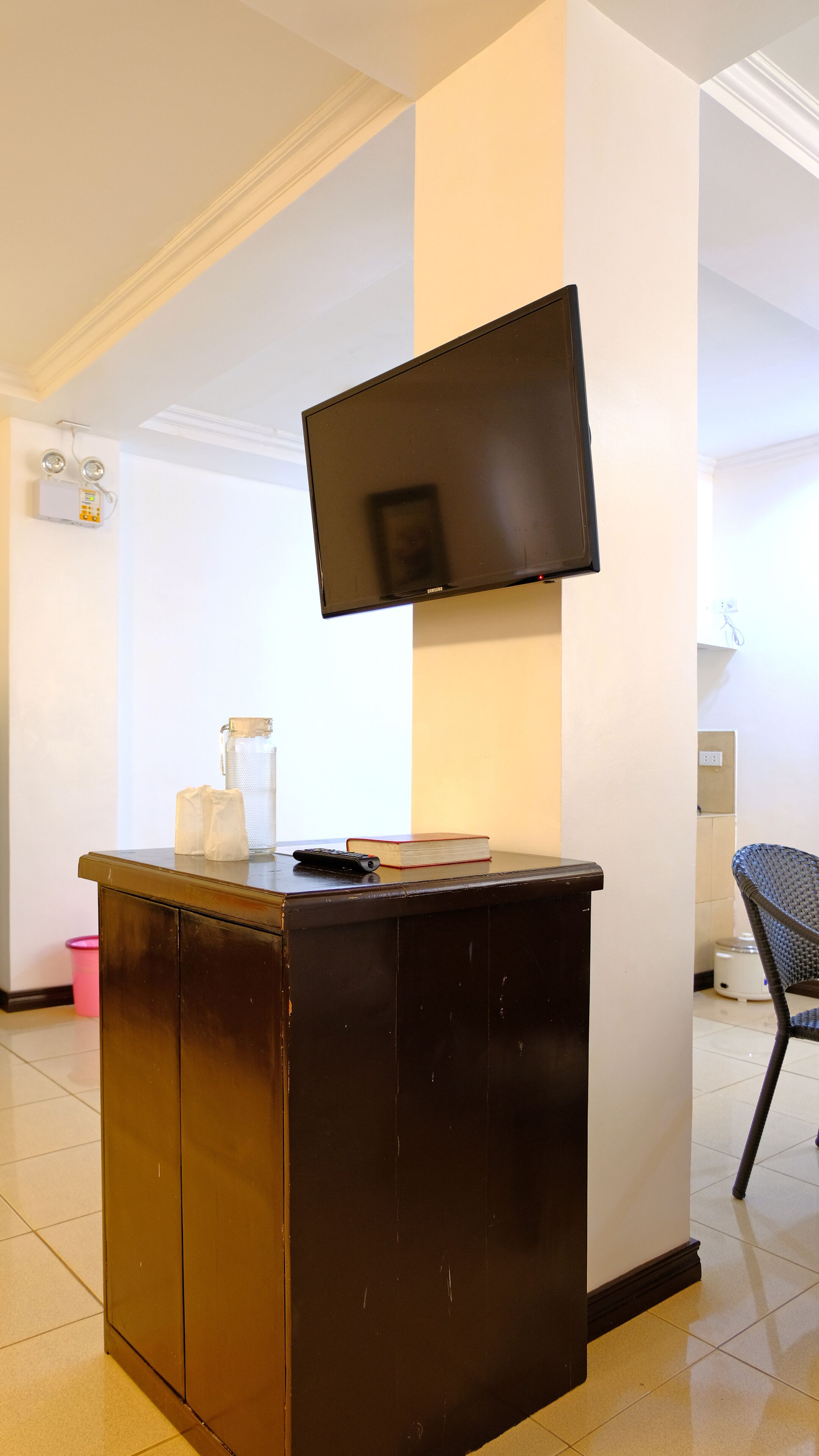 A room with a flat-screen TV mounted on the wall, a small wooden desk with a book and remote control, a trash bin, and a chair.