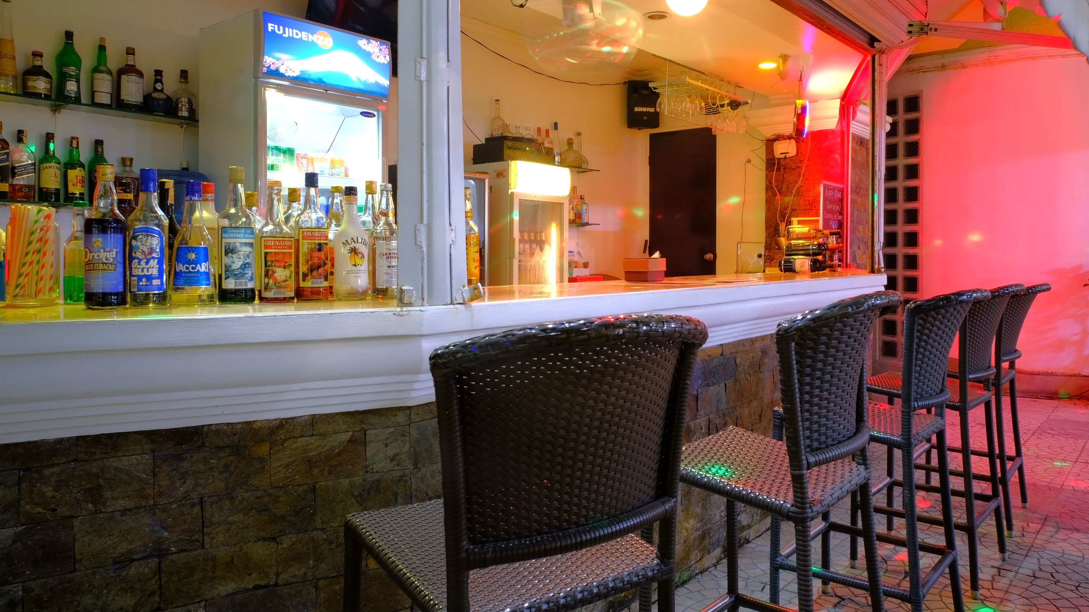 Un espace bar avec un assortiment de bouteilles d’alcool, un présentoir réfrigéré et des tabourets de bar en osier.