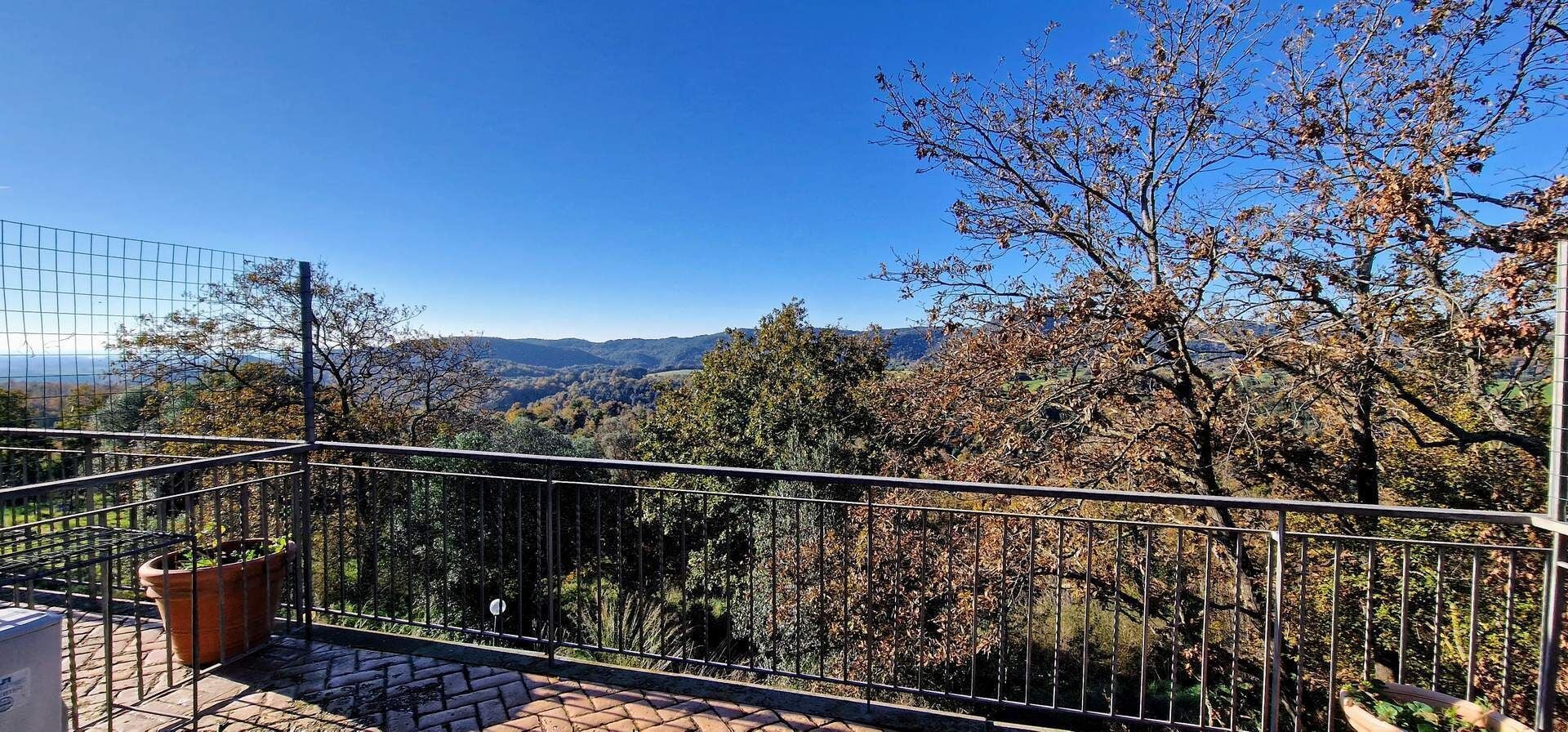 Un balcón con barandilla de metal que da a un paisaje montañoso con árboles y cielo despejado.