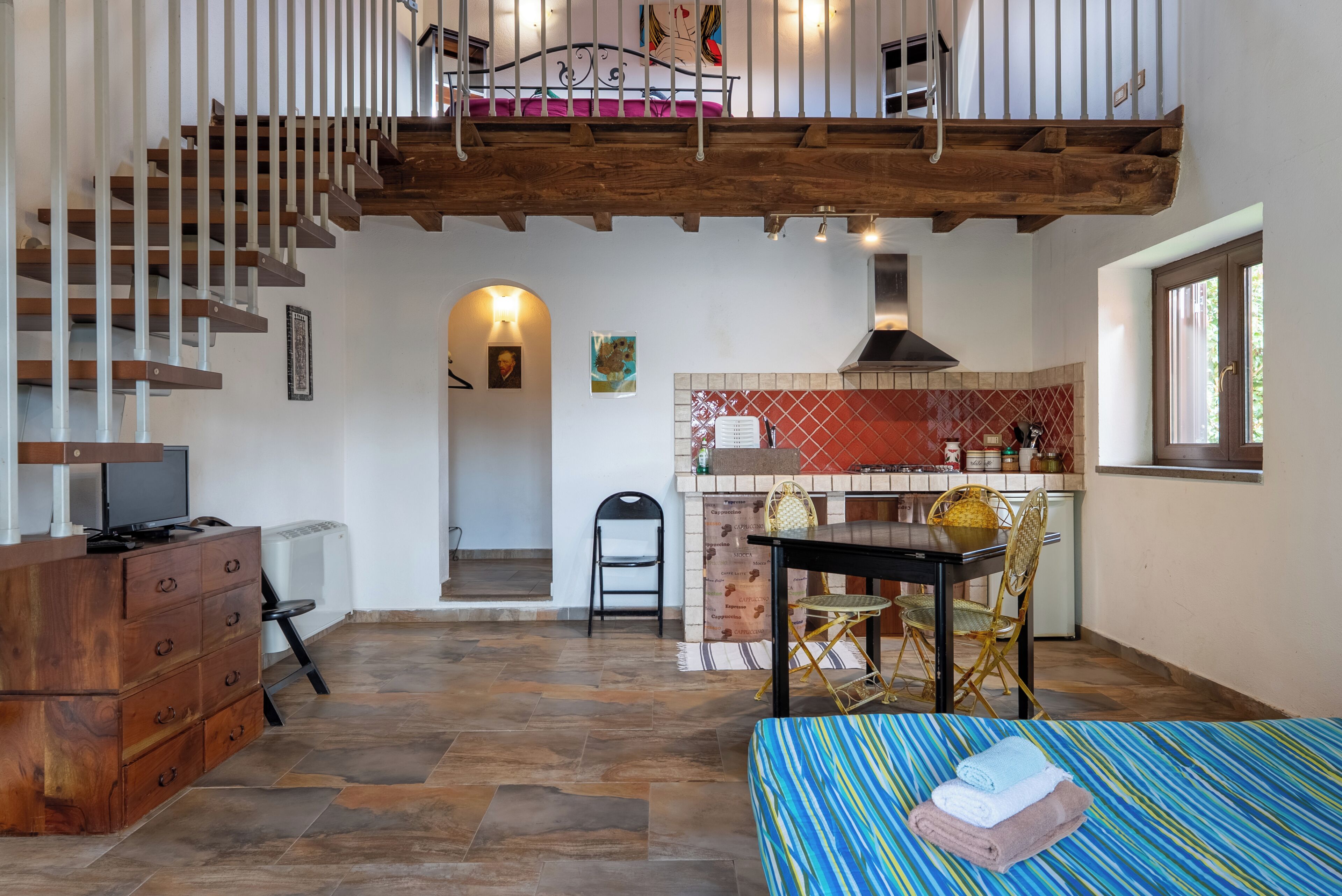 A spacious room with a wooden staircase, a dining area with a table and chairs, a kitchen with a red tiled backsplash, and a bed with striped bedding.