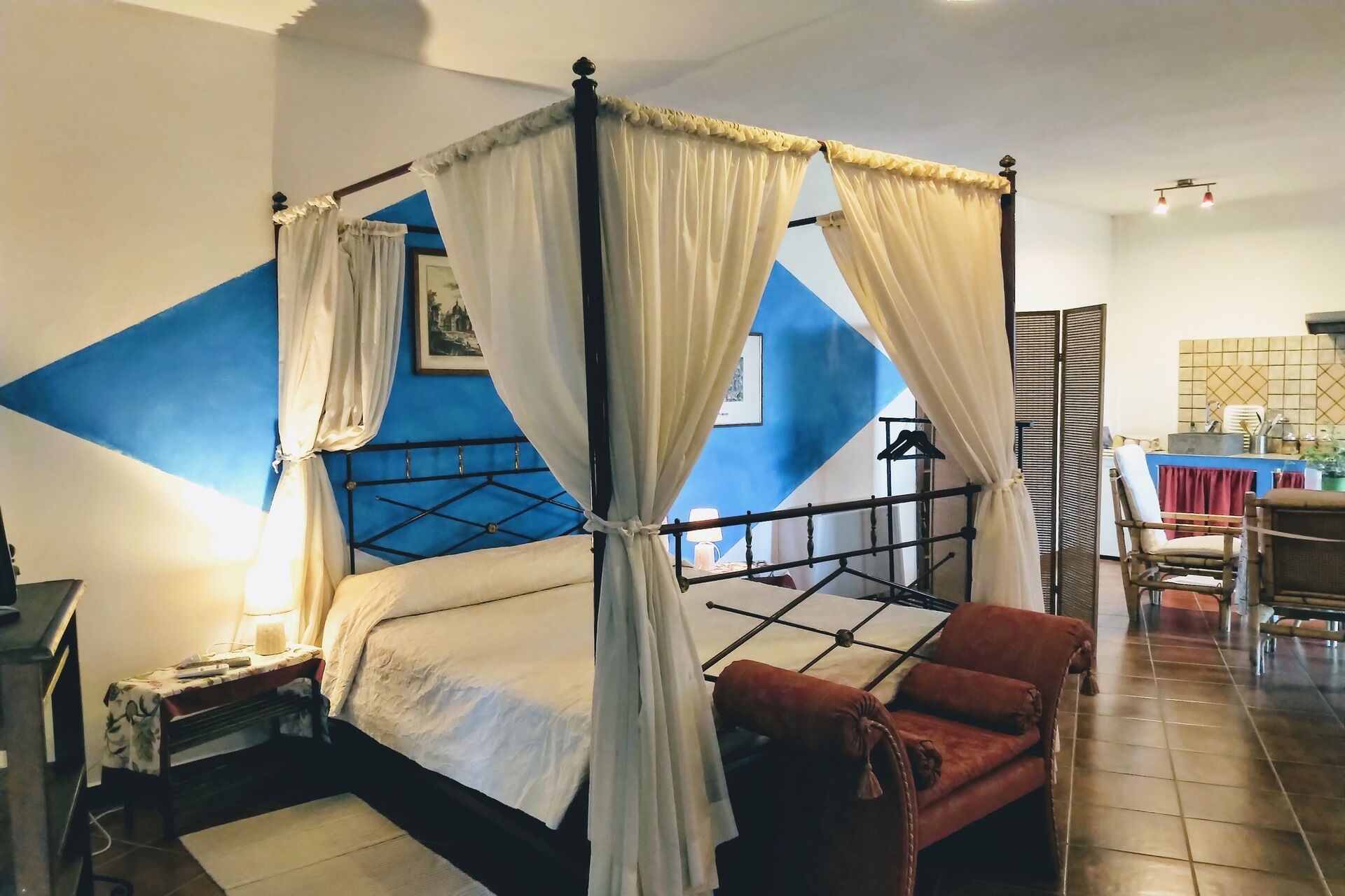 Un dormitorio con una cama con dosel, un escritorio con lámpara, una silla y vistas al área de la cocina.