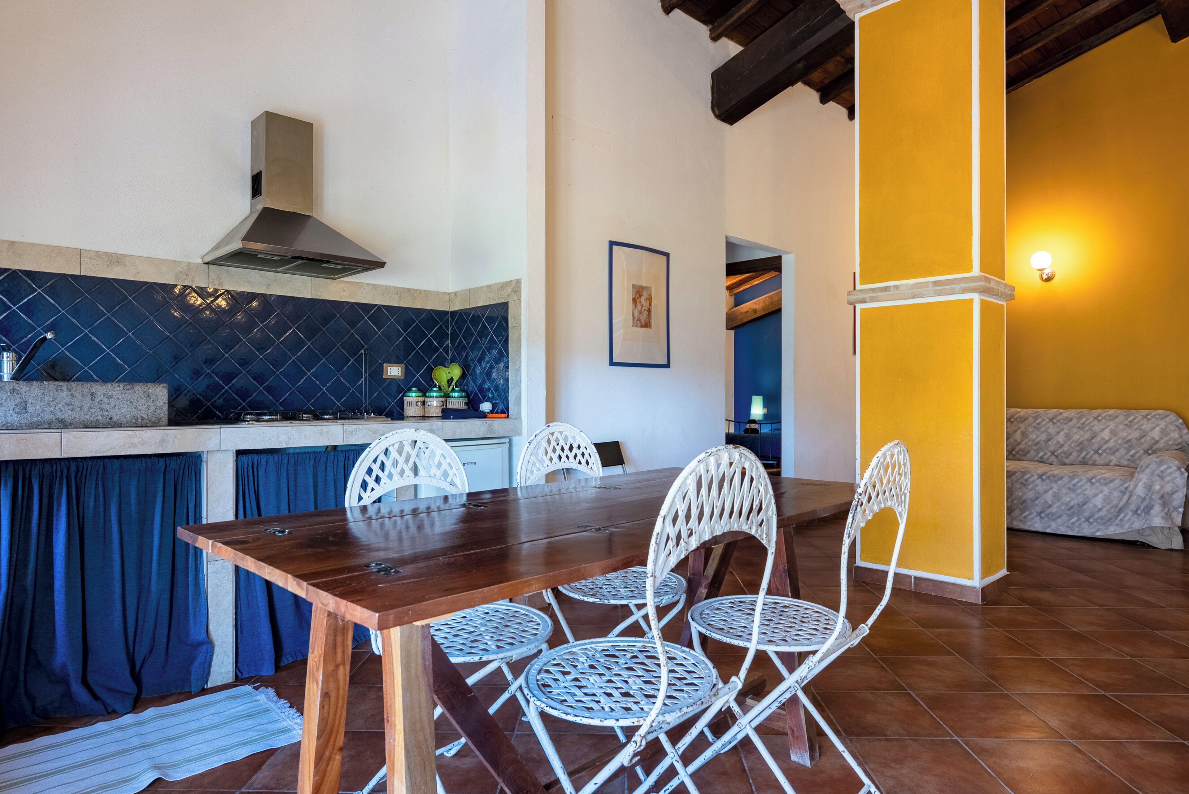 A dining area with a wooden table and white chairs, a kitchen with blue tiles, and a yellow wall.