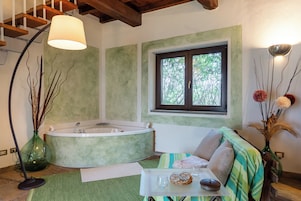 A room with a large bathtub, a window with a view of trees, a sofa with cushions, and a tray with glasses and a bowl.