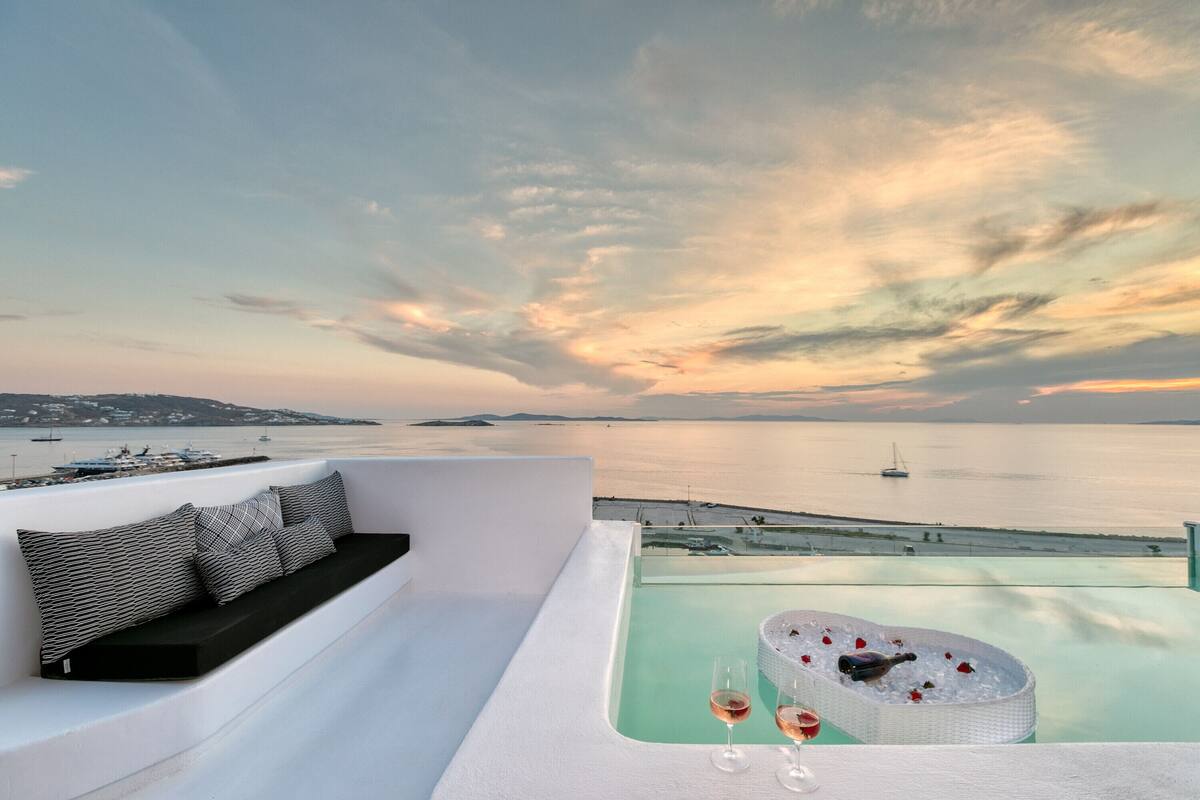 Una piscina infinita en la azotea con borde de cristal, un sofá blanco con cojines estampados y dos copas de vino tinto junto a la piscina.