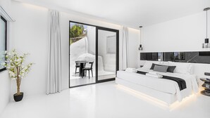 Un dormitorio moderno con una cama grande, una zona de comedor con mesa y sillas, y un ventanal corredero que da a un espacio exterior.