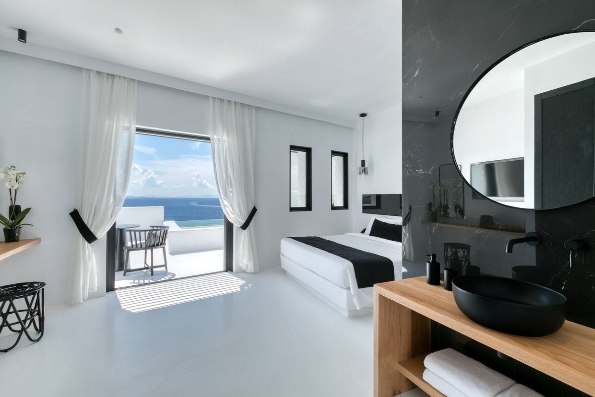 Una habitación de hotel moderna con una cama grande, un espejo redondo, un lavamanos negro y vistas al océano.