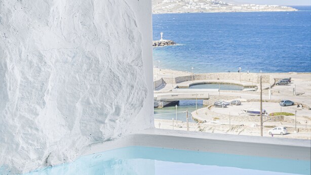 Vista desde una habitación con pared blanca y ventana que da a una zona costera con piscina y muelle.