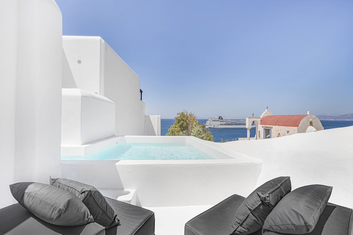 Una piscina infinita blanca con cojines grises, con vistas a un mar azul cristalino y un crucero.