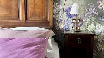 A bedroom with a wooden headboard, a bed with white and purple bedding, a nightstand with a lamp, and floral wallpaper.