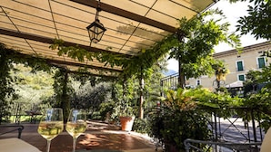 Un patio al aire libre con dos copas de vino sobre una mesa, rodeado de follaje y un edificio al fondo.