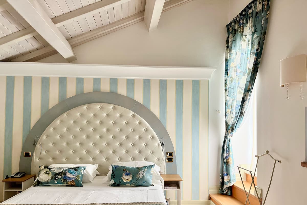 Ein Schlafzimmer mit einer karrierten Kopfteile, gestreifter Tapete, einem Bett mit floralen Kissen, einem Nachttisch mit Lampe und einem Fenster mit Vorhängen.