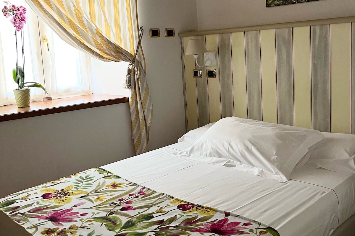 Ein Schlafzimmer mit einem Bett, einem Fenster mit Vorhängen, einer Zimmerpflanze und einem Bild an der Wand.