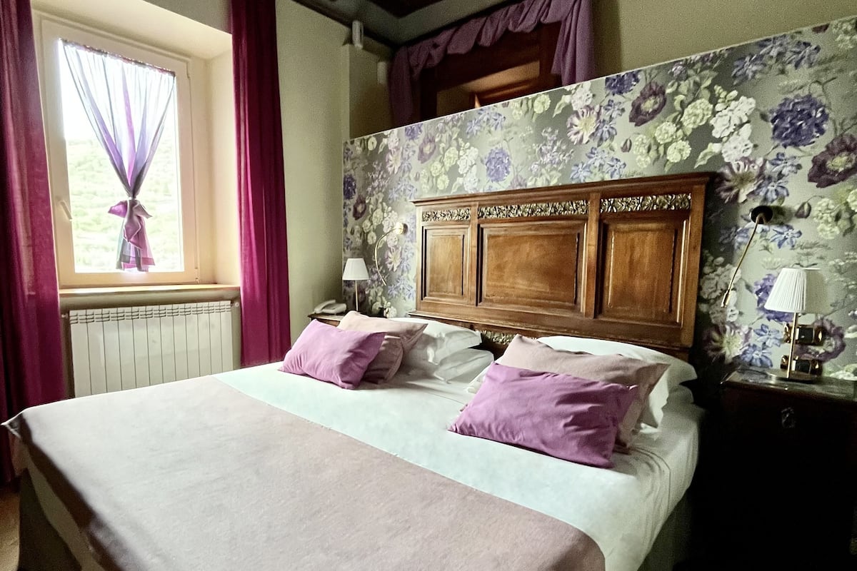 Ein Schlafzimmer mit einem großen Bett, einem Fenster mit Vorhängen, einem Wandpaneel mit Blumenmuster am Bettfuß und einem Nachttisch mit Lampe.