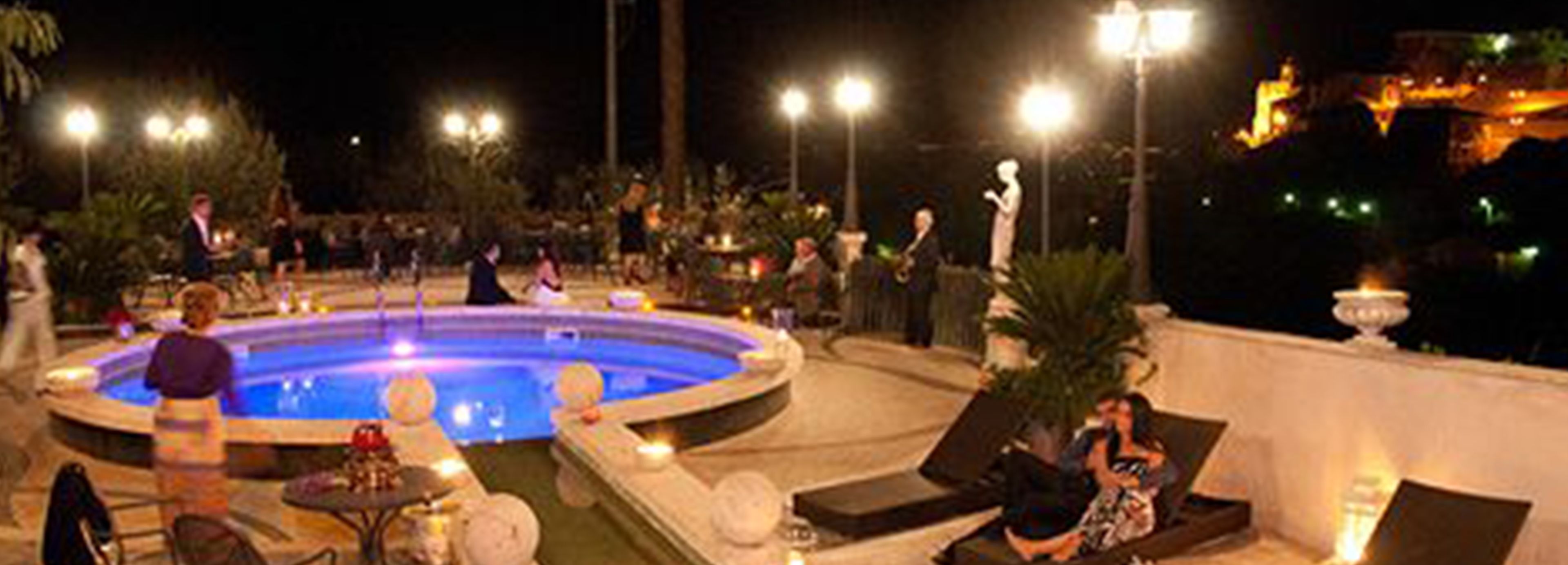 Uma área de piscina ao ar livre à noite, com espreguiçadeiras e mesas, cercada por caminhos bem iluminados e plantas decorativas.