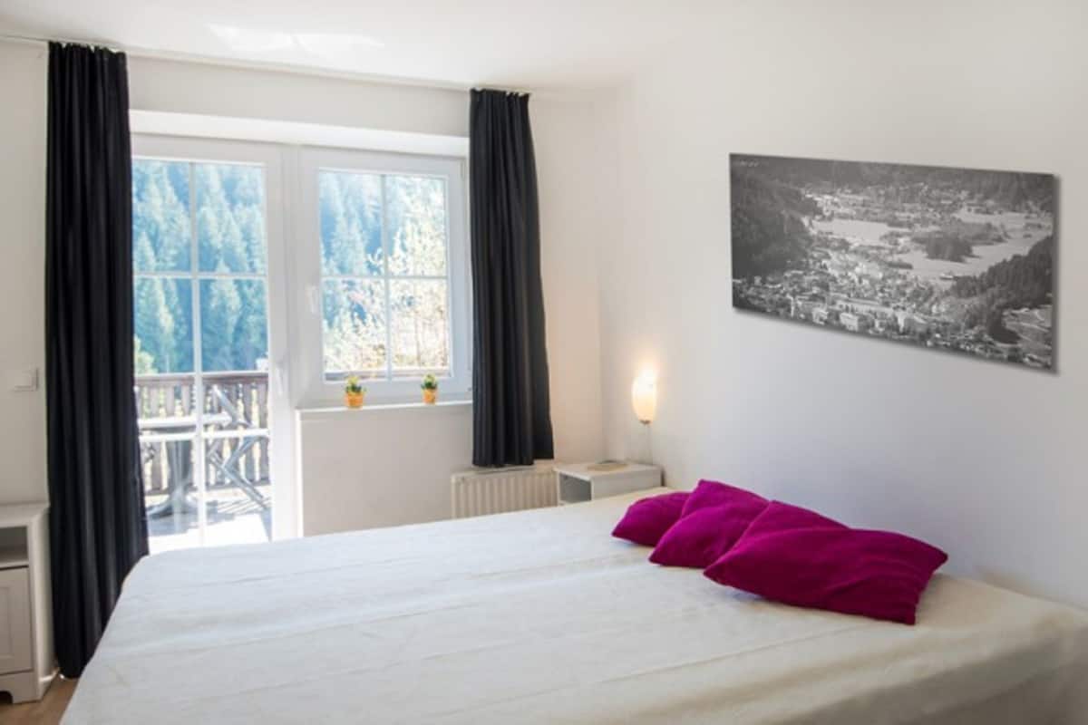 A bedroom with a bed, a window with curtains, a radiator, a nightstand, a wall-mounted light, and a black and white photograph.
