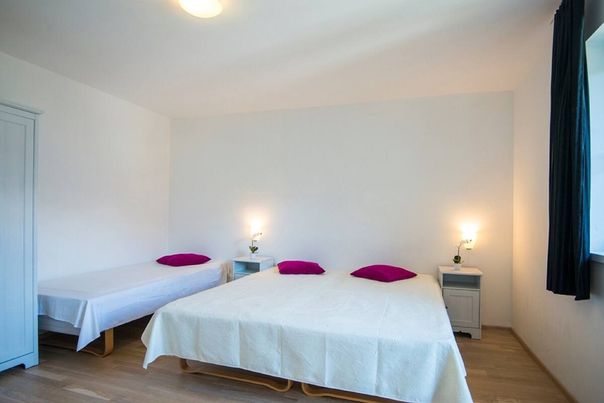A bedroom with two single beds and a larger bed, each with white linens and pink pillows. There are bedside tables with lamps.