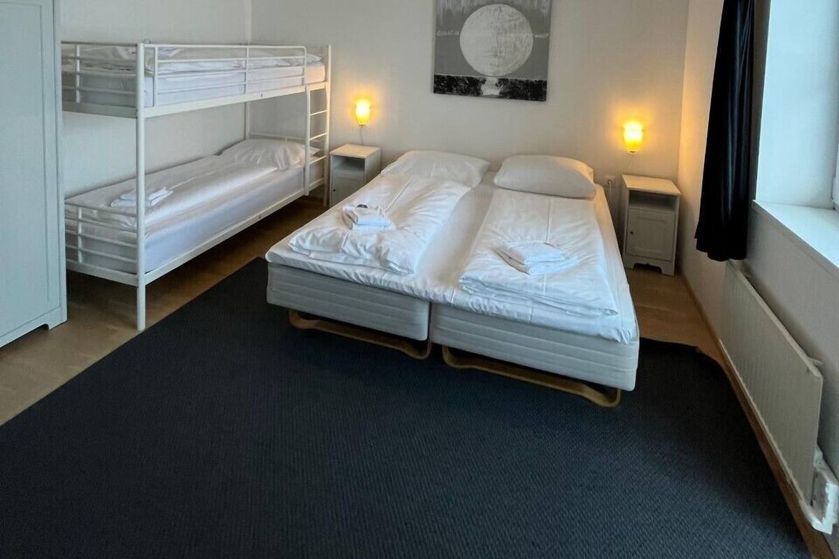 A room with bunk beds, a single bed, a wall-mounted artwork, bedside lamps, and a window with curtains.