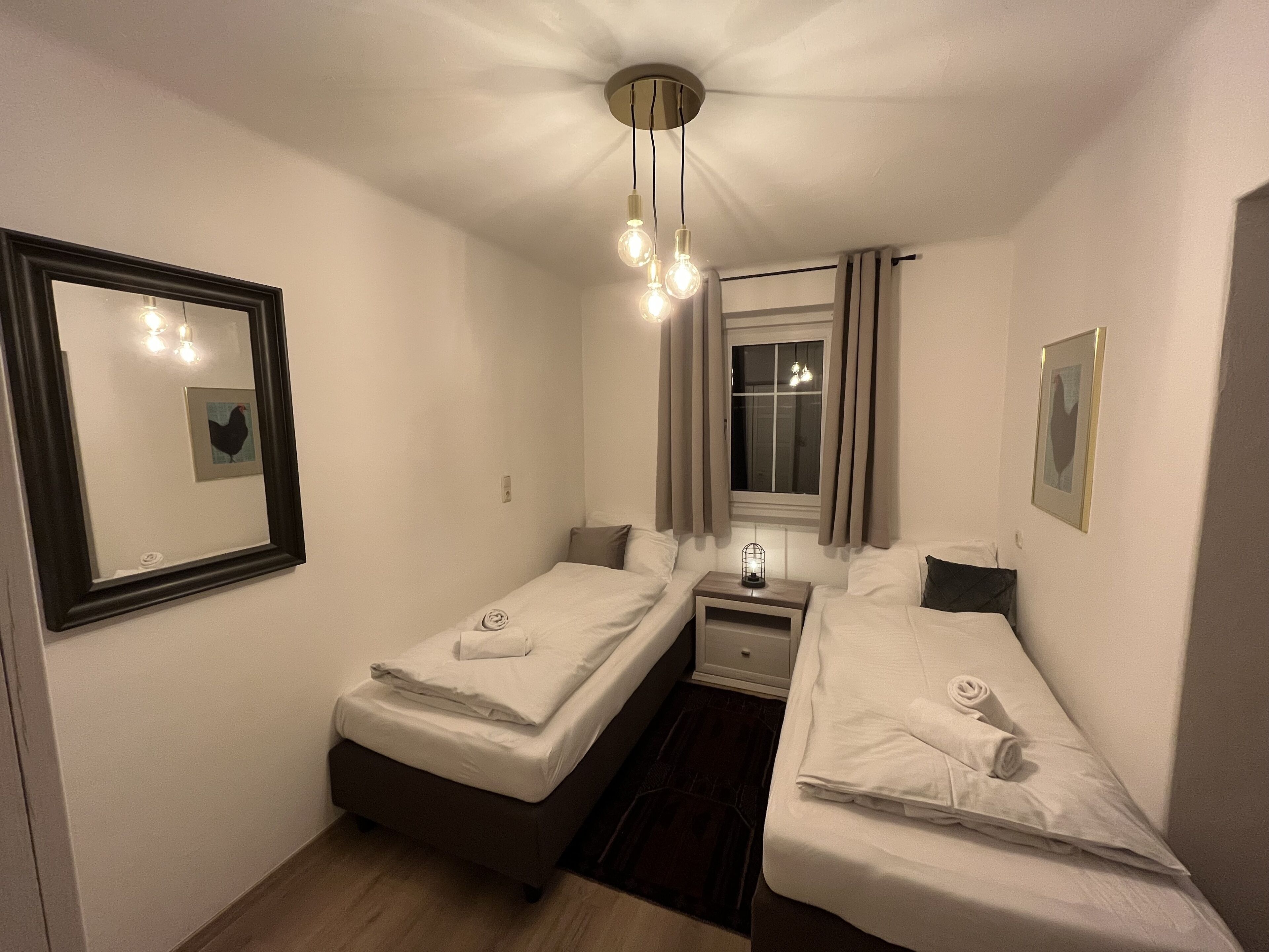Un dormitorio pequeño y bien iluminado con dos camas individuales, una mesita de noche con lámpara, una ventana con cortinas y un espejo en la pared.