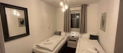 Un dormitorio pequeño y bien iluminado con dos camas individuales, una mesita de noche con lámpara, una ventana con cortinas y un espejo en la pared.