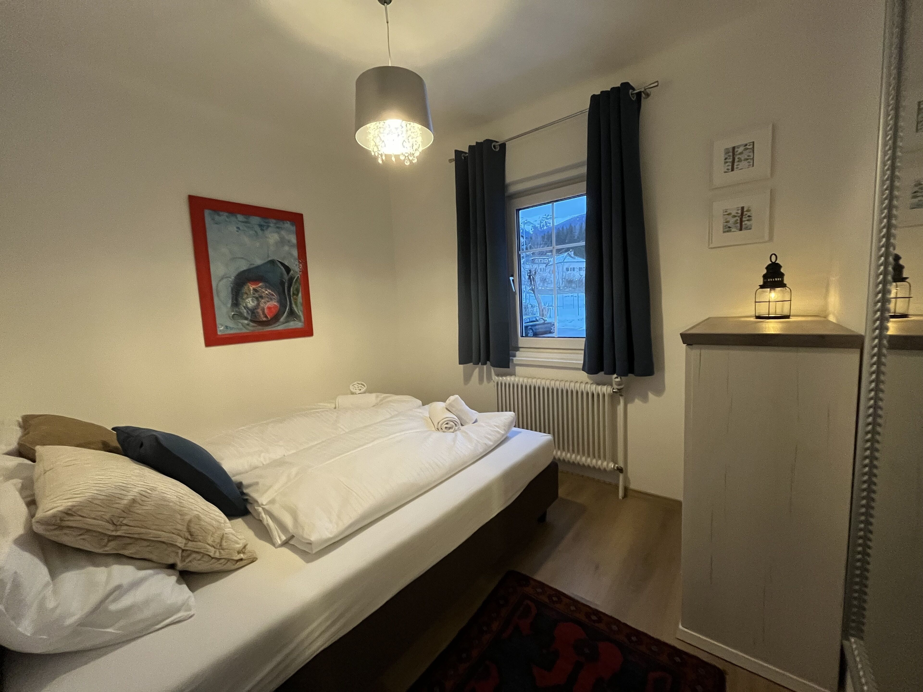 Een slaapkamer met een enkel bed, een radiator, een raam met gordijnen, een schilderij aan de muur en een klein tafeltje met een lantaarn.