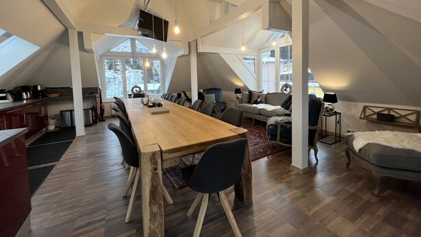 Un ampio loft con soffitto alto, un lungo tavolo da pranzo in legno e un’accogliente zona salotto con divano e poltrone.