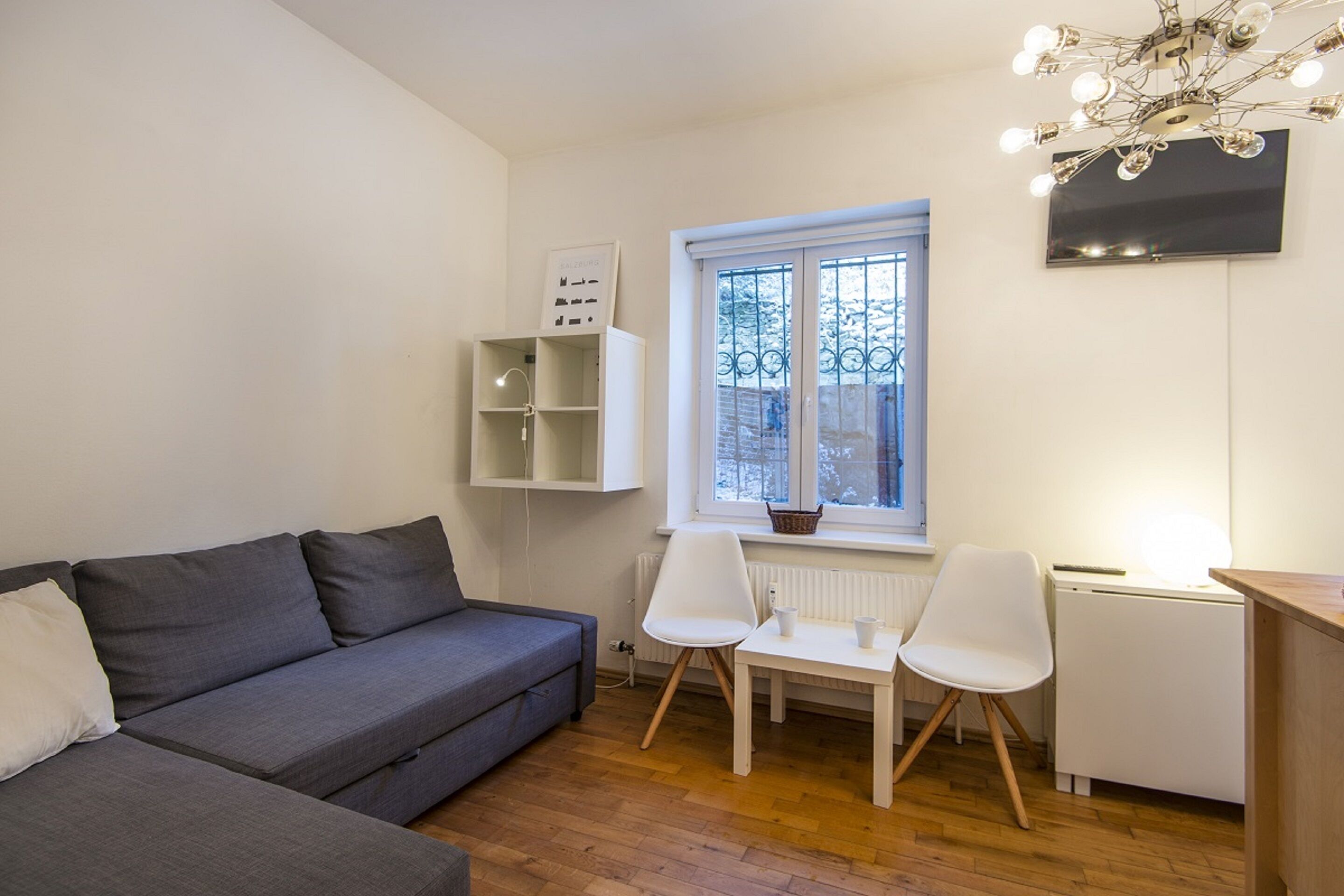 A modern living room with a grey sofa, white chairs, a small table, a white cabinet, and a TV mounted on the wall.