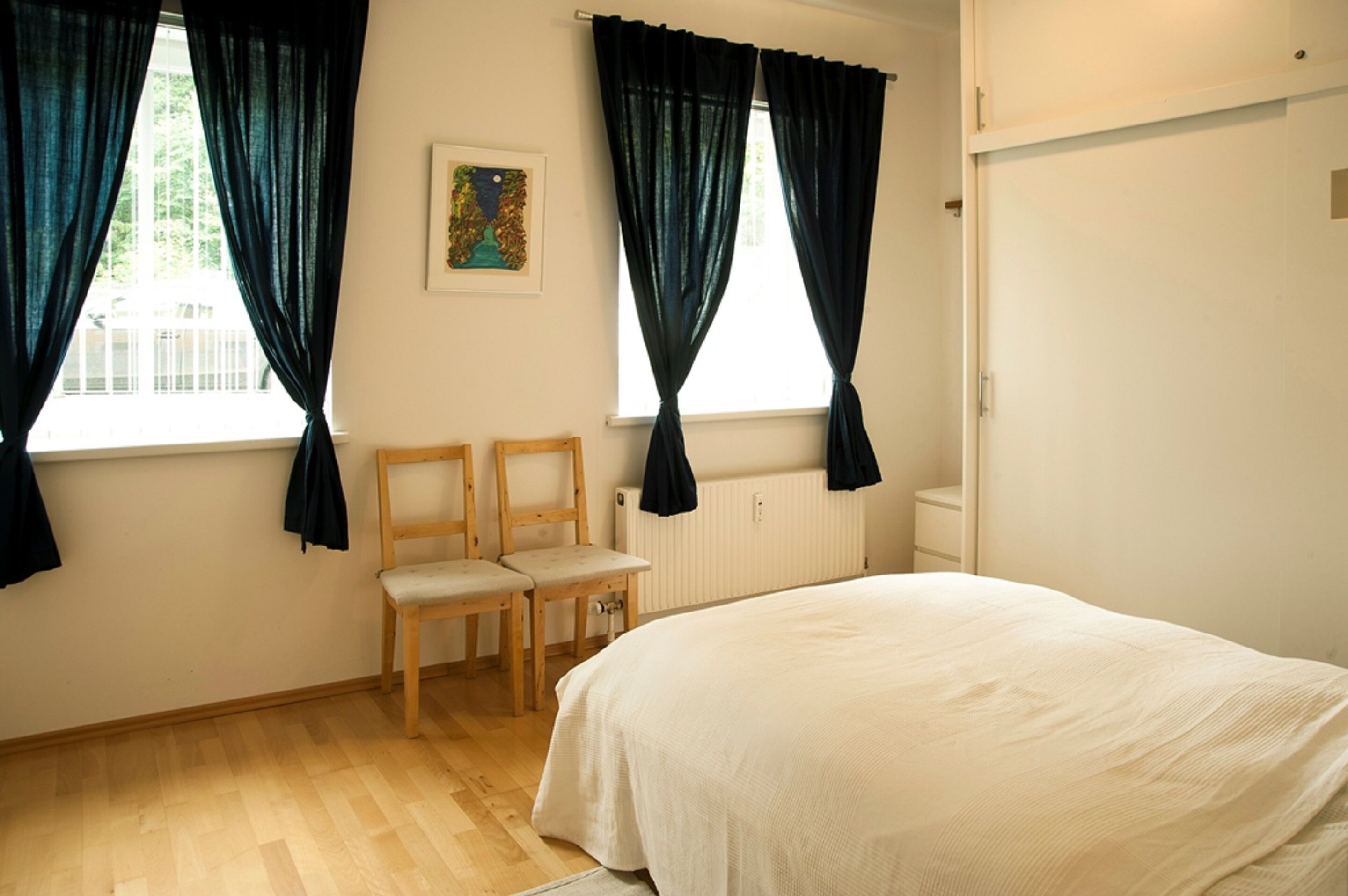 Une chambre à coucher avec un lit, deux chaises, un radiateur et une fenêtre avec des rideaux.