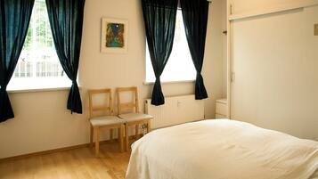 Un dormitorio con una cama, dos sillas, un radiador y una ventana con cortinas.