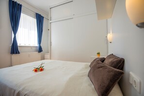 A hotel room with a bed, blue curtains, a small potted plant, and a wall-mounted light fixture.