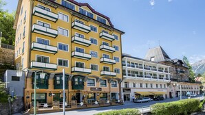Un hotel de varios pisos con balcones, un restaurante en la planta baja y una montaña al fondo.