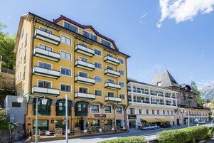 Un hotel de varios pisos con balcones, un restaurante en la planta baja y una montaña al fondo.