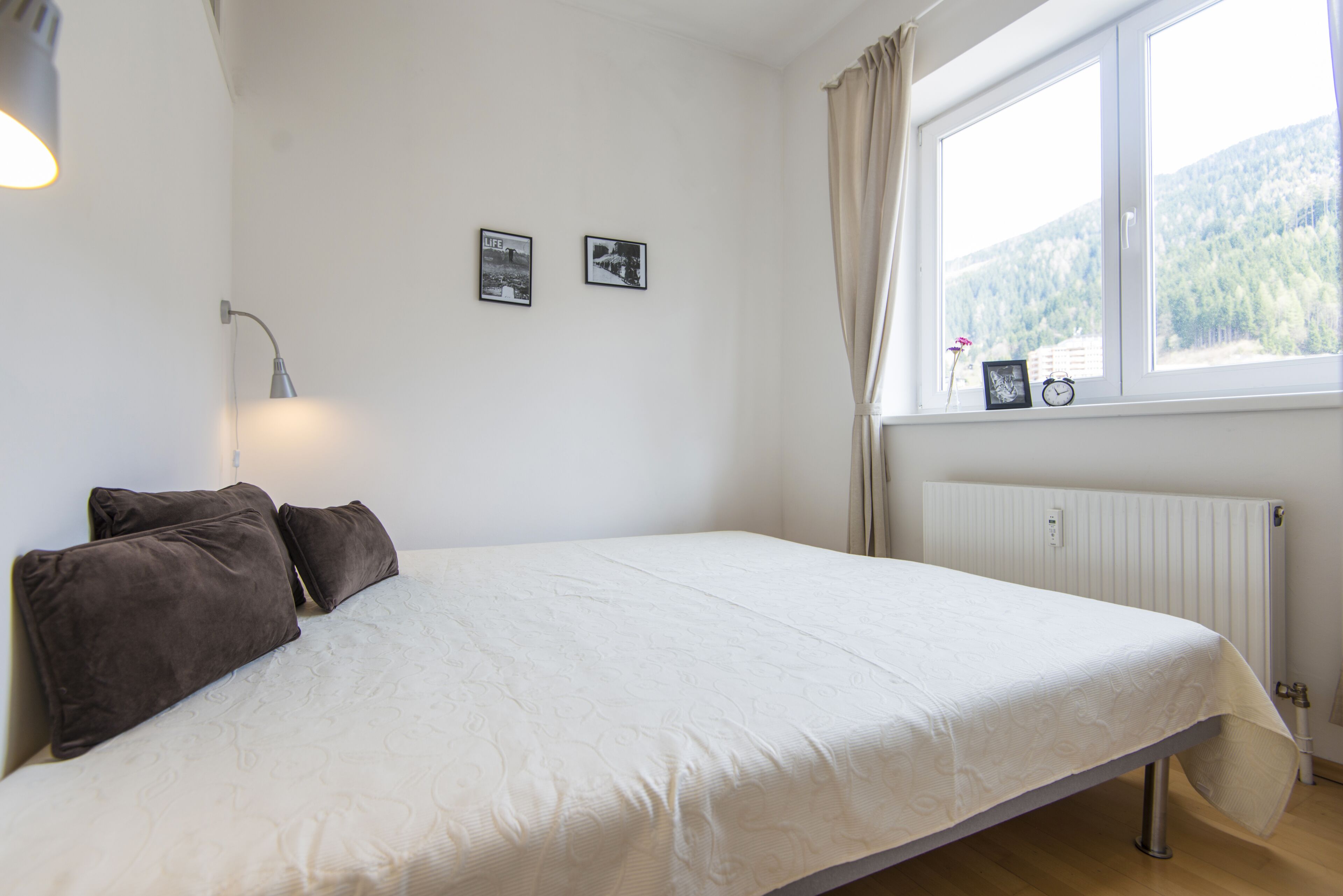 A bedroom with a bed, a window with curtains, a radiator, and two framed pictures on the wall.