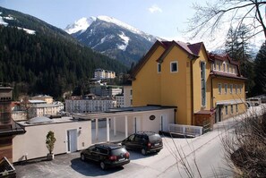 A yellow building with a red roof, a parking area with two cars, and a mountain backdrop.