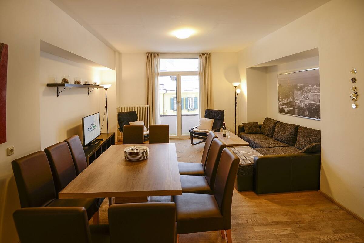 A modern living room with a wooden dining table, comfortable seating, a large window with curtains, a sofa, and a wall-mounted TV.