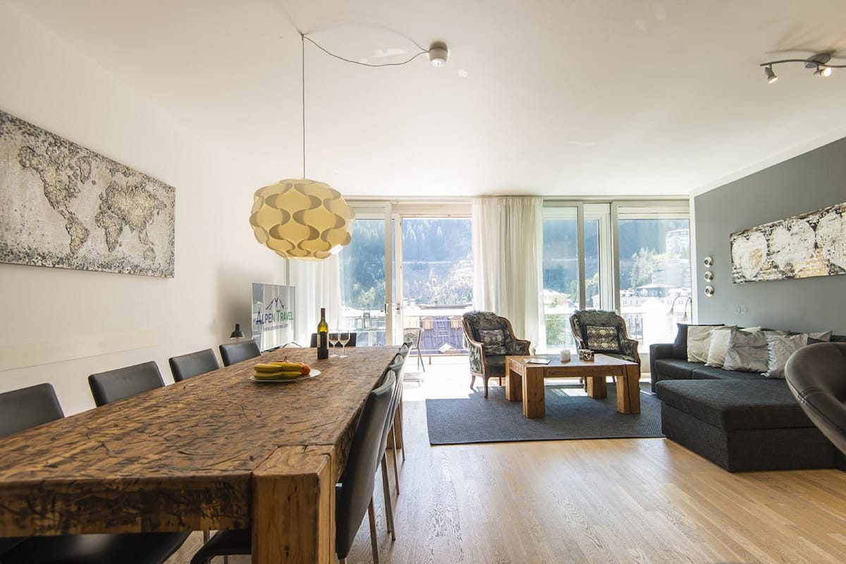 A spacious living area with a wooden dining table, a sofa, and a coffee table. There are chairs around the table, and a large window provides a view of the city.