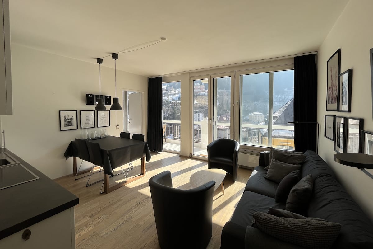 A modern living room with a large window, a dining area, and a kitchen.