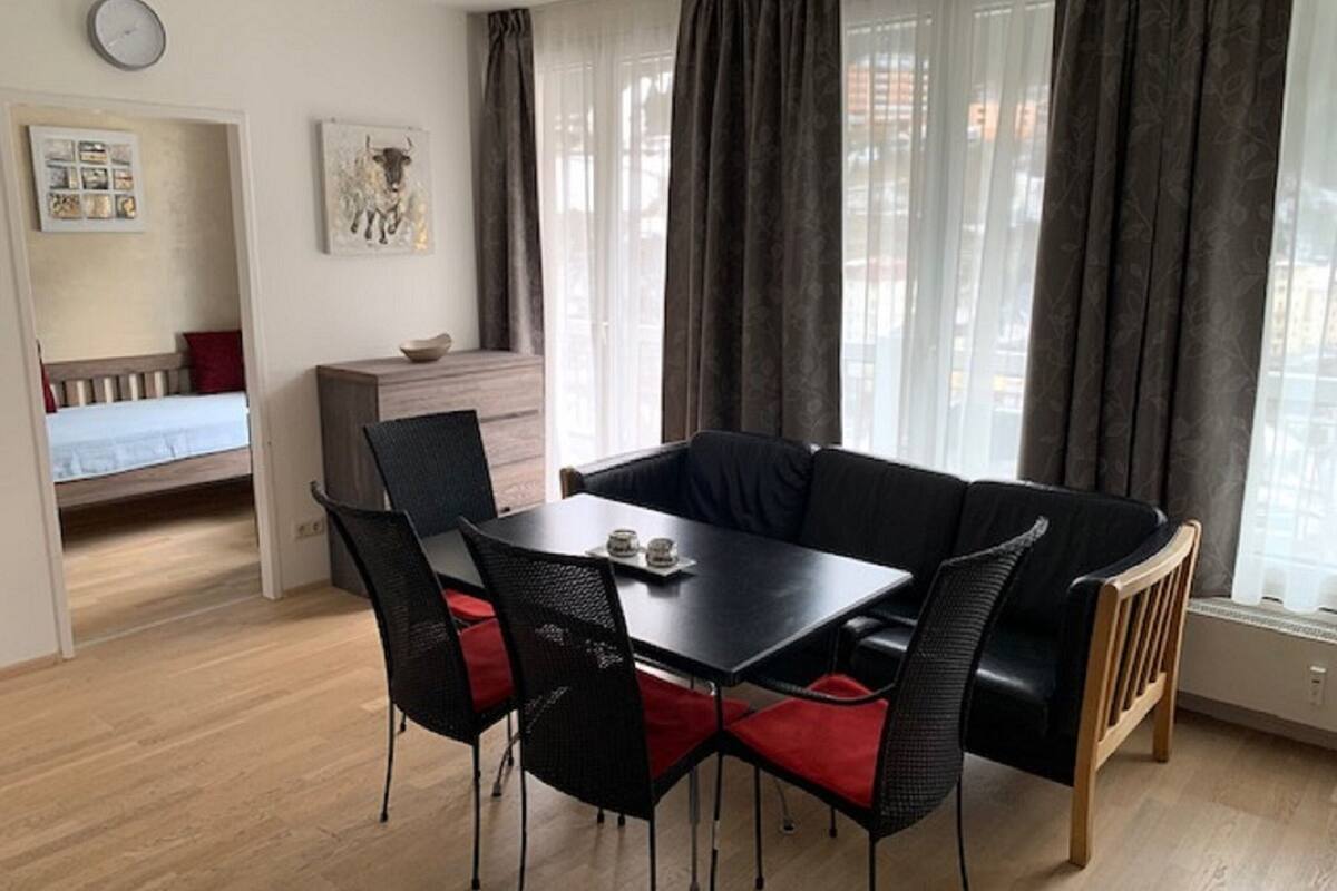A dining area with a black table and chairs, a wooden bench, a small kitchenette, and a bed in the background.