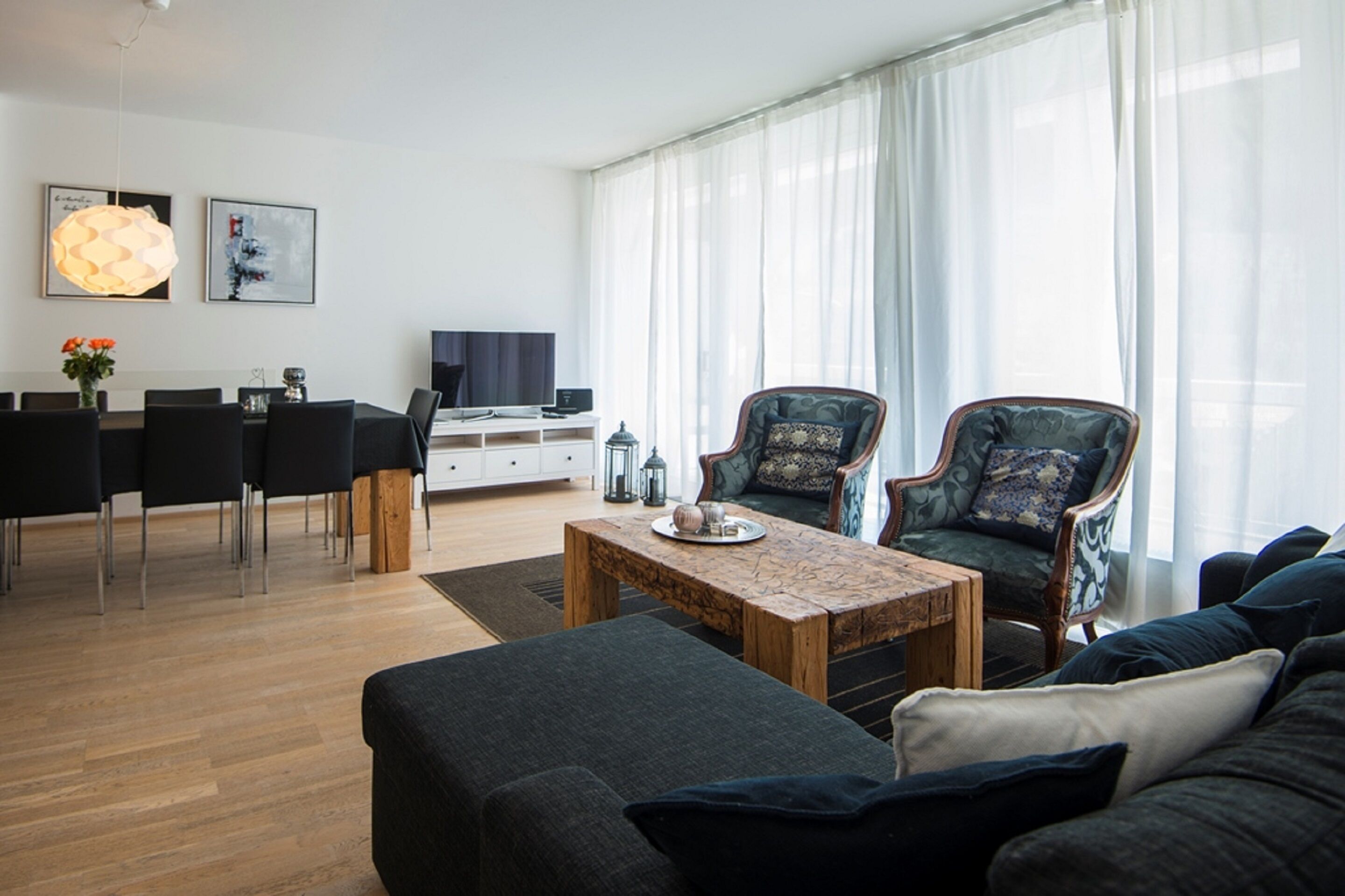 Una sala moderna con un sofá, dos sillones, una mesa de centro, una zona de comedor con sillas y una mesa, un televisor y obras de arte en la pared.