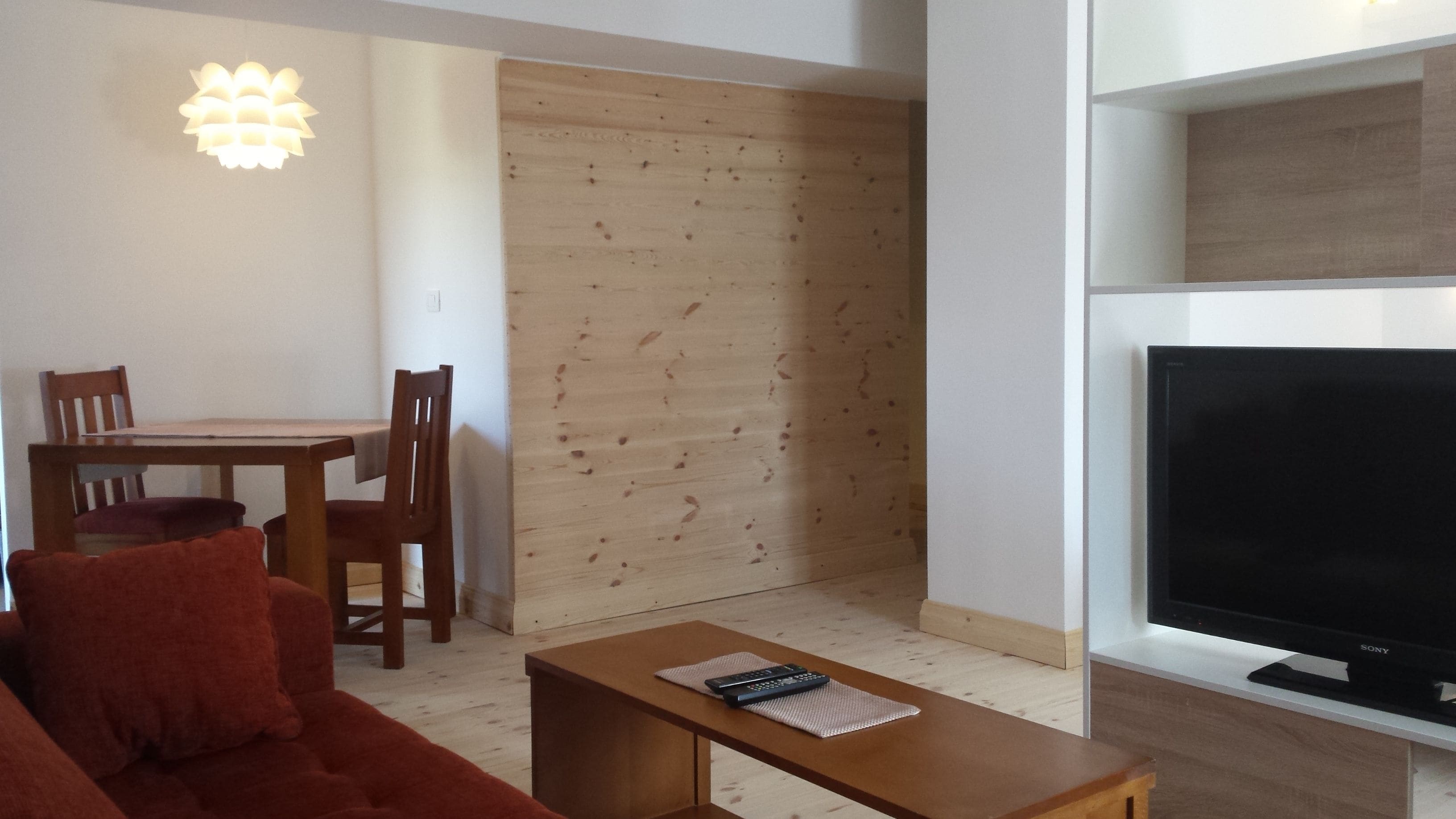 A living room with a sofa, a coffee table, a TV, a wooden dining table with chairs, and a wall with a wooden panel.