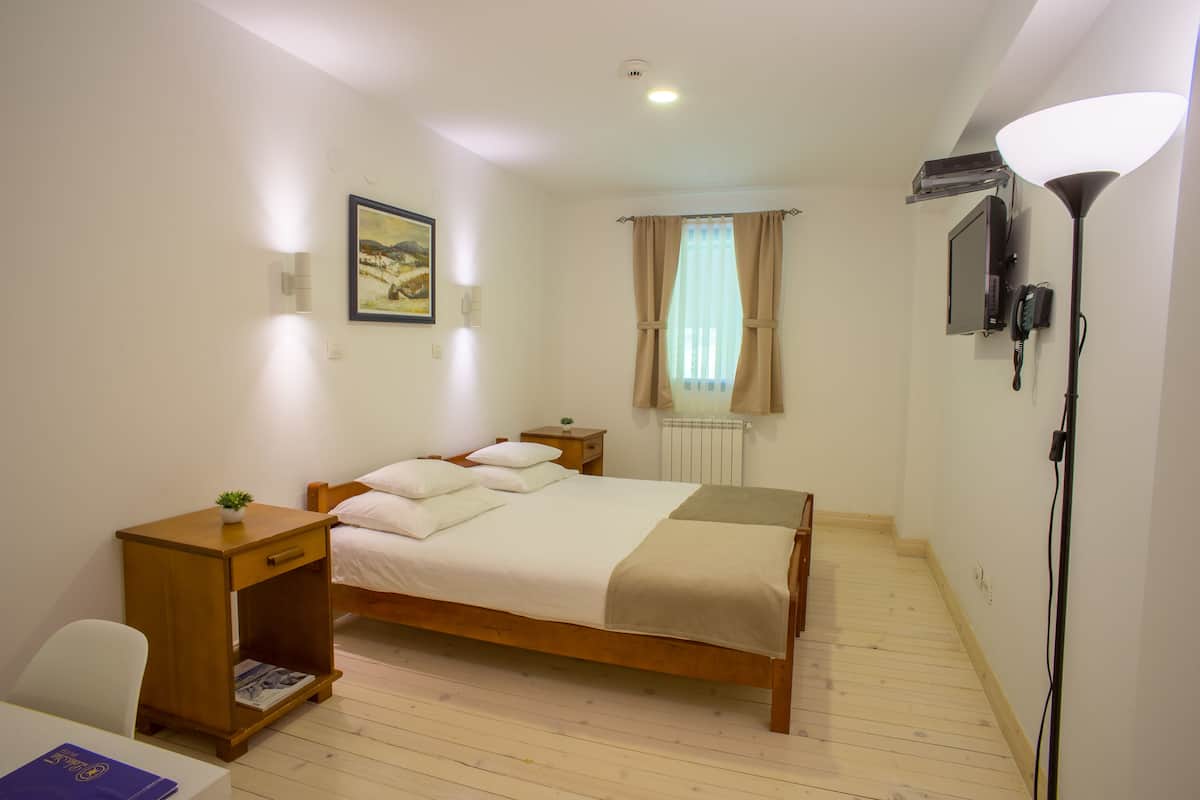 A bedroom with a wooden bed, bedside tables, a lamp, a radiator, and a wall-mounted TV.