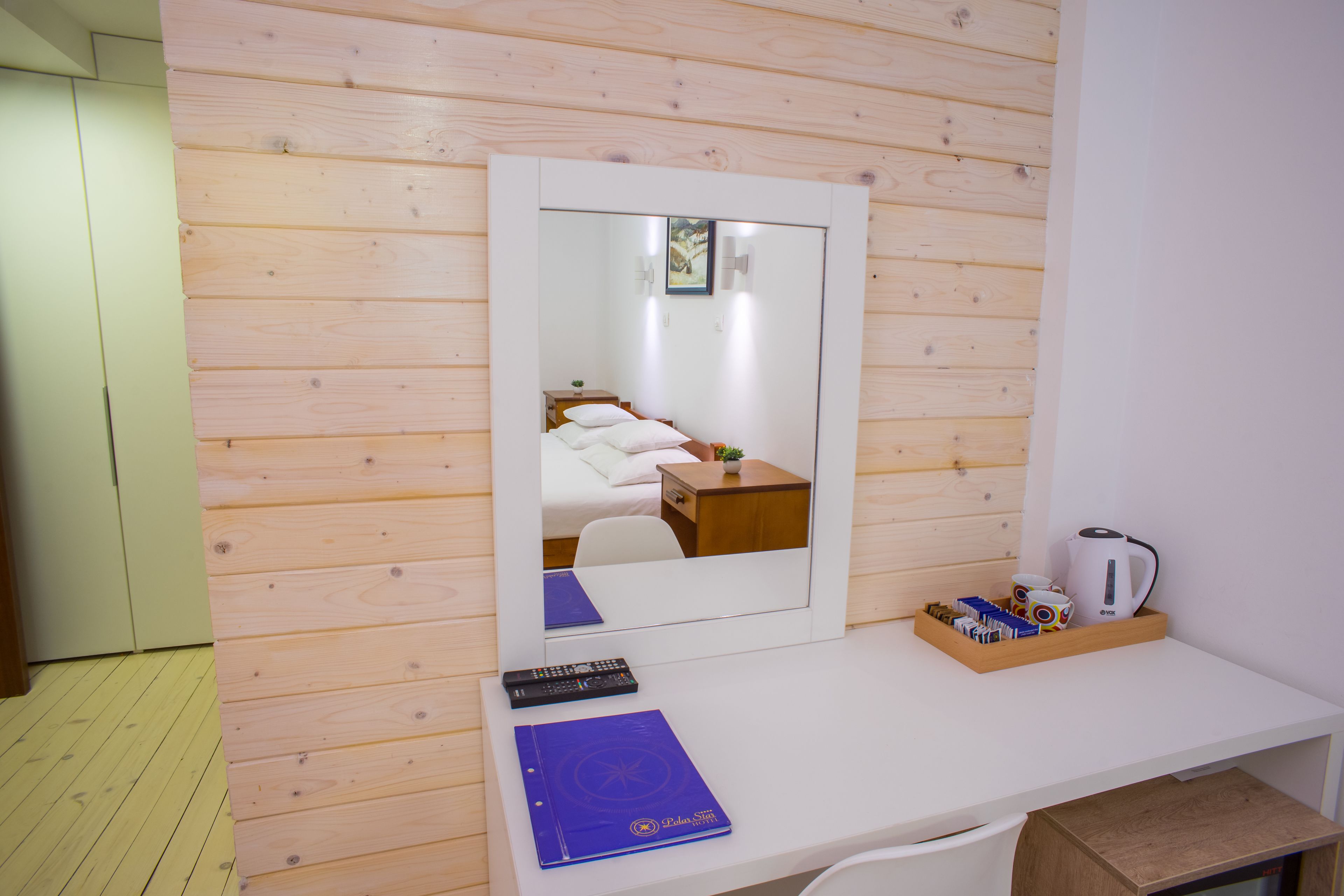 A room with a wooden wall, a white desk with a mirror, a bed, a nightstand, a small table, and a tray with cups and a kettle.
