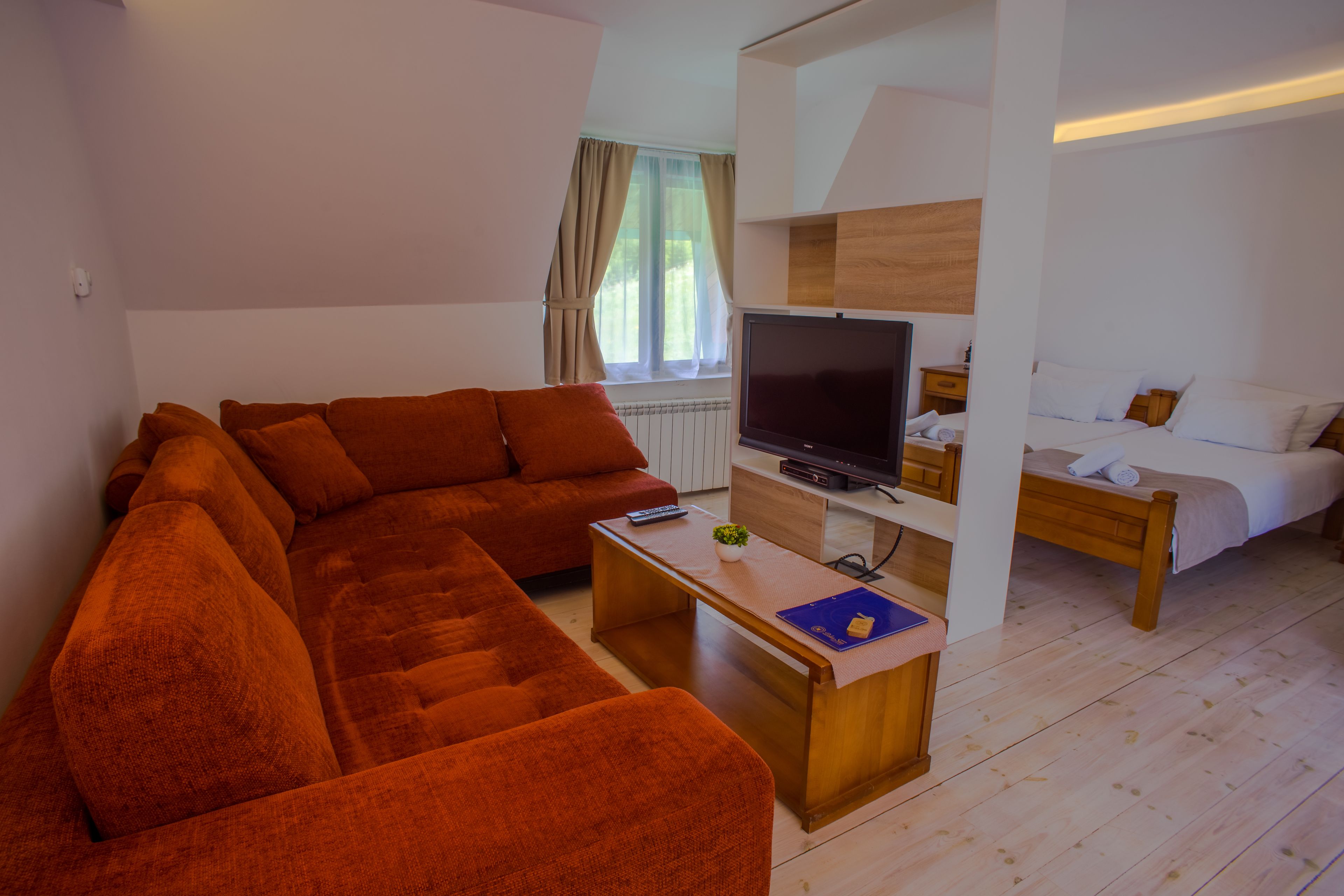 A modern living room with a sectional sofa, a flat-screen TV, a small table with a remote and a book, and a separate bedroom with a bed and a nightstand.